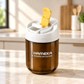A HAMEKA amber brown water bottle with a white lid and a yellow flip-up straw, set against a solid beige background.